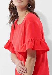 Rotes, kurzärmliges Oberteil mit rüschenartigen Kanten, aus glattem Stoff gefertigt. Verfügt über einen runden Ausschnitt und eine lockere Passform. Elegante Creolen ergänzen den Look.