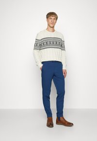 Cream cable-knit sweater with black geometric patterns, paired with blue tailored pants and brown leather ankle boots.