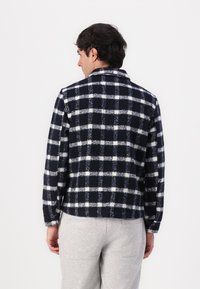 Marineblå og hvit rutete jakke i ullblandingsstoff, med lange ermer, krage og cropped design. Brukt over grå bukser.