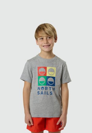 Ragazzo biondo sorridente che indossa una maglietta grigia "North Sails" con loghi colorati di barche a vela e pantaloncini arancioni vivaci, in piedi su uno sfondo semplice.