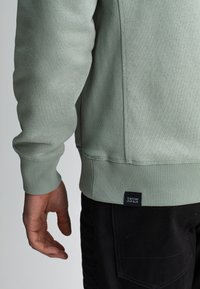 Lichtgroene sweatshirt met ribgebreide manchetten, zijnaad en zoom. Etiket zegt "BUTCHER OF BLUE." Draagt met zwarte broek, waarbij een hand ernaast rust.