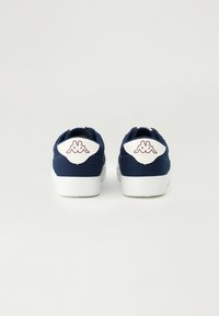 Baskets en toile bleu marine avec un tirant blanc portant un logo, semelle en caoutchouc et accents rouges au niveau du col et des oeillets.