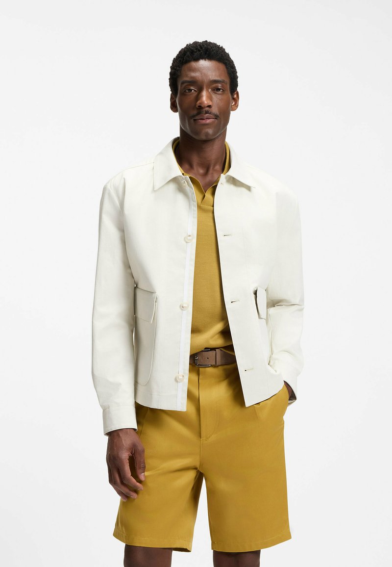 Homme portant une veste blanche boutonnée par-dessus un polo moutarde avec un short moutarde assorti et une ceinture marron sur fond blanc.