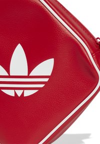 Pochette in pelle sintetica rossa con logo Adidas trefoil bianco e bordatura zippata bianca coordinata, caratterizzata da una superficie testurizzata e forma angolare.