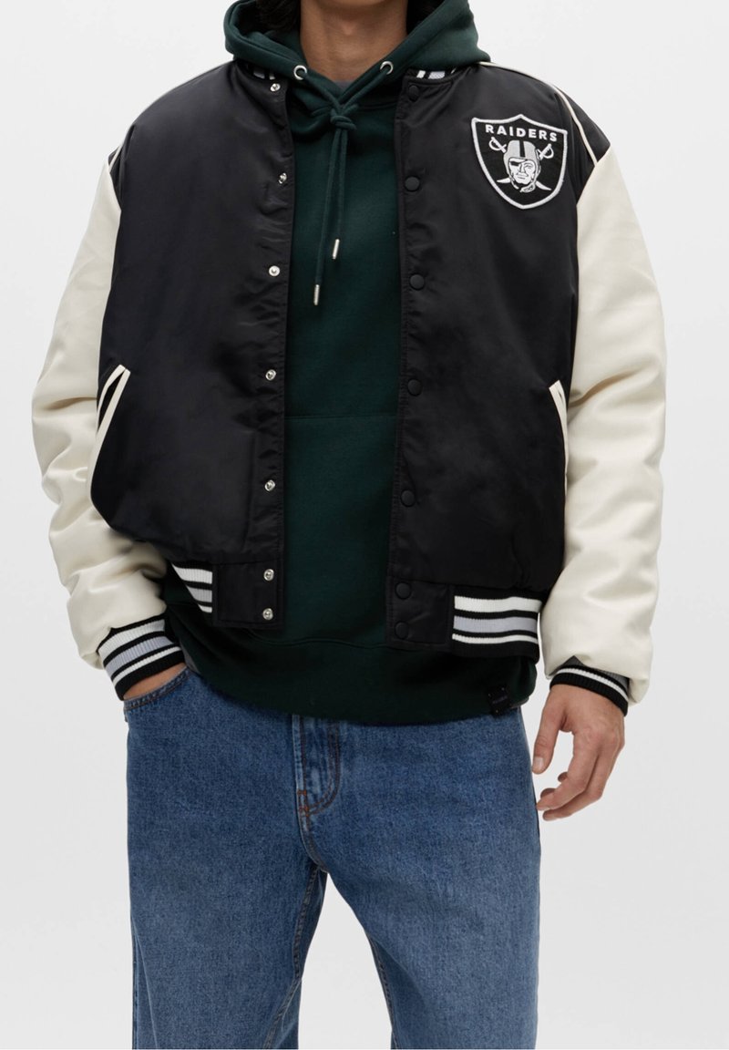 Zwarte en crèmekleurige varsityjack met een Raider's logo, knoopsluiting, geribbelde kraag en gestreepte manchetten, gedragen over een groene hoodie en jeans.