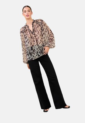 Kobieta ubrana w długą, prześwitującą bluzkę w panterkę, czarne szerokie spodnie oraz czarne sandały z odkrytymi palcami, stojąca z jedną ręką na biodrze.