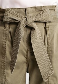 Närbild av khakifärgad byxor med ett texturerat tygbälte knutet i en rosett vid midjan.