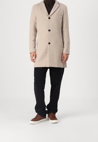 Beige wool coat with a notched collar, three buttons, one chest pocket, side pockets, and black buttons. Paired with black trousers and brown shoes.