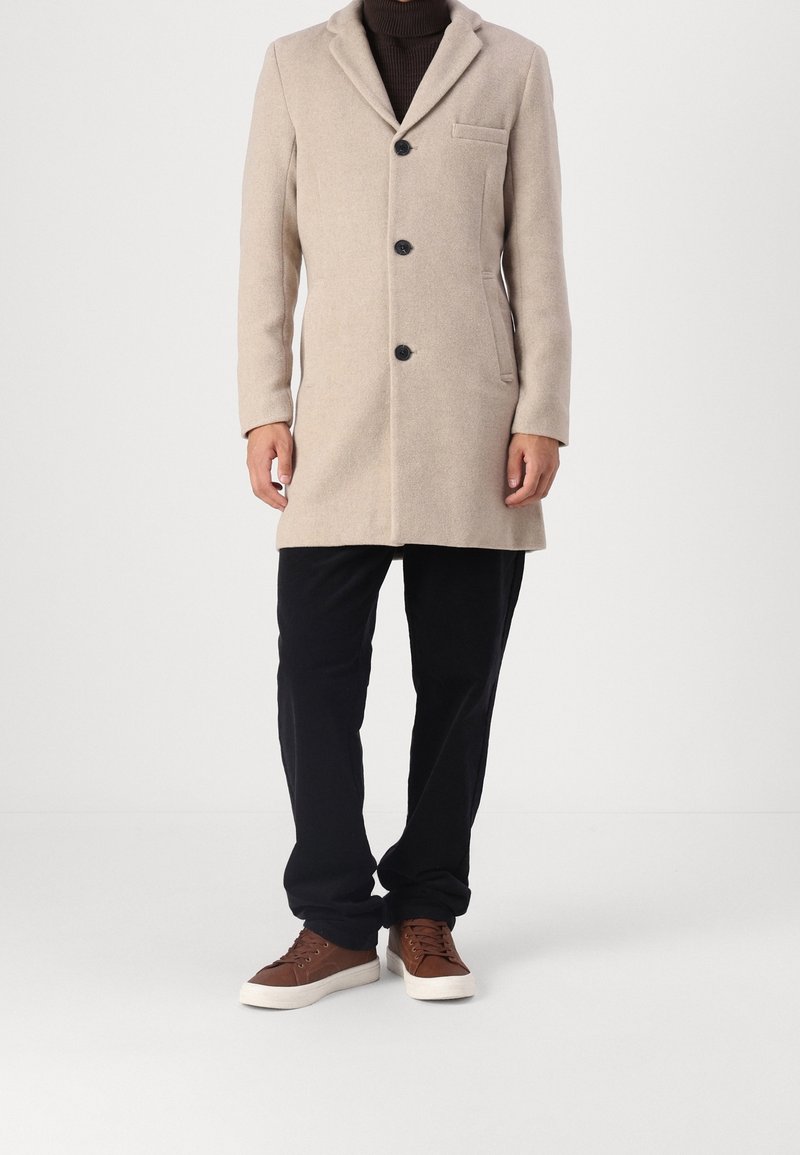 Beige wool coat with a notched collar, three buttons, one chest pocket, side pockets, and black buttons. Paired with black trousers and brown shoes.