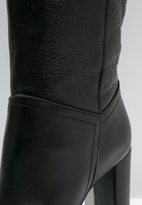 Schwarze Leder-Stiefelette mit hohem Absatz, strukturiertem Finish, Nahtdetails und einem glatten Akzent am Knöchel.