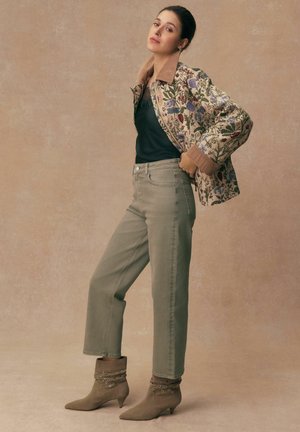 Frau steht mit einer Hand in der Tasche, trägt eine jacke mit Blumenmuster, ein schwarzes Oberteil, olivgrüne Hosen und beige knöchelhohe Stiefel mit Ösen.