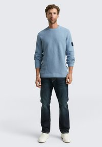 Lichtblauwe gebreide trui met een textuurpatroon, ronde hals en geribde mouwen, gecombineerd met donkerblauwe jeans en witte sneakers.