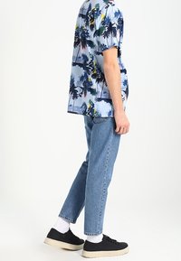 Kurzarmhemd mit einem blauen Palmenmuster, kombiniert mit hellblauen geraden Jeans und schwarzen Sneakers, die weiße Socken tragen.