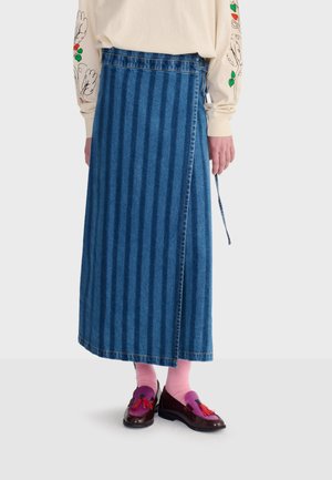 Osoba w niebieskiej dżinsowej spódnicy z zakładką w paski, kremowej bluzce z długim rękawem z kwiatowym wzorem na rękawach, różowych skarpetkach i fioletowych mokasynach z chwostami.