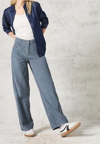 Kvinna klädd i lösa blå byxor, vita sneakers, vit linne och en oknäppt mörkblå skjorta, stående mot en ljus strukturerad vägg.