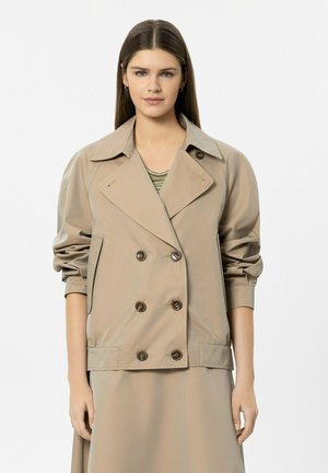 Young woman with straight brown hair wearing a beige double-breasted jacket and matching skirt on a white background.