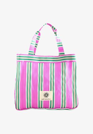 Pink tote bag with vertical green, white, and beige stripes and fabric handles, featuring a "self studios" logo patch with a sun design.