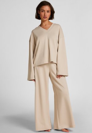 Jonge vrouw in een beige, loszittend V-hals topje en wijde broekspijpen, staande op blote voeten tegen een effen achtergrond.