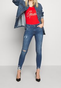 Chaqueta de mezclilla con lavado azul, jeans desgastados con desgastes y dobladillo deshilachado, combinados con una blusa roja ajustada con el logo plateado de "Guess" y tacones negros.