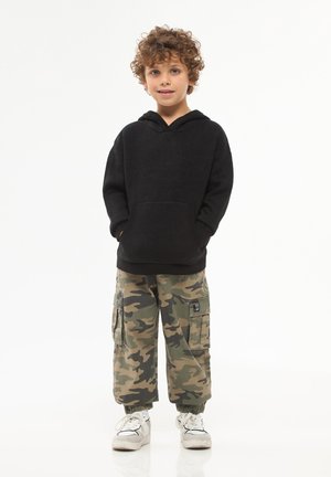 Felpa nera con cappuccio e tasca frontale, abbinata a pantaloni cargo mimetici con tasche laterali e polsini elastici, indossata con sneakers bianche.
