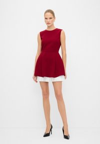 Robe sans manches rouge avec jupe plissée blanche, ourlet évasé, fabriquée dans un tissu lisse ; portée avec des talons hauts noirs et des accessoires minimalistes.