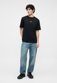 Jaunas vyras stovi, dėvėdamas juodą trumpomis rankovėmis marškinėlį, šviesiai mėlynas laisvas džinsas ir juodus sportinius batelius, prie paprasto šviesaus fono.