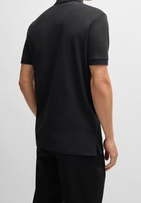 Musta poolopaita, jossa on sileä kangas, lyhyet hihat ja klassinen kaulus. Sivulenkit helman kohdalla liikkuvuuden helpottamiseksi.