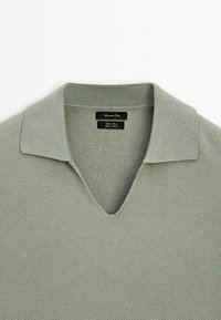 Polo en maille texturée vert sauge clair avec un col en V profond et une étiquette de marque noire à l'intérieur du col.