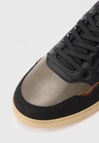 Primo piano di una punta di sneaker con pelle grigia testurizzata, sovrapposizioni in suede e pelle nere, lacci neri e una suola in gomma beige.