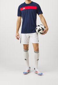 Camiseta deportiva a rayas en azul marino y rojo, pantalones cortos blancos, calcetines blancos con detalles en negro, sosteniendo un balón de fútbol en blanco y negro.