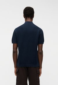 Homme debout, dos à la caméra, portant un polo à manches courtes bleu marine et un short cargo foncé, sur un fond neutre.