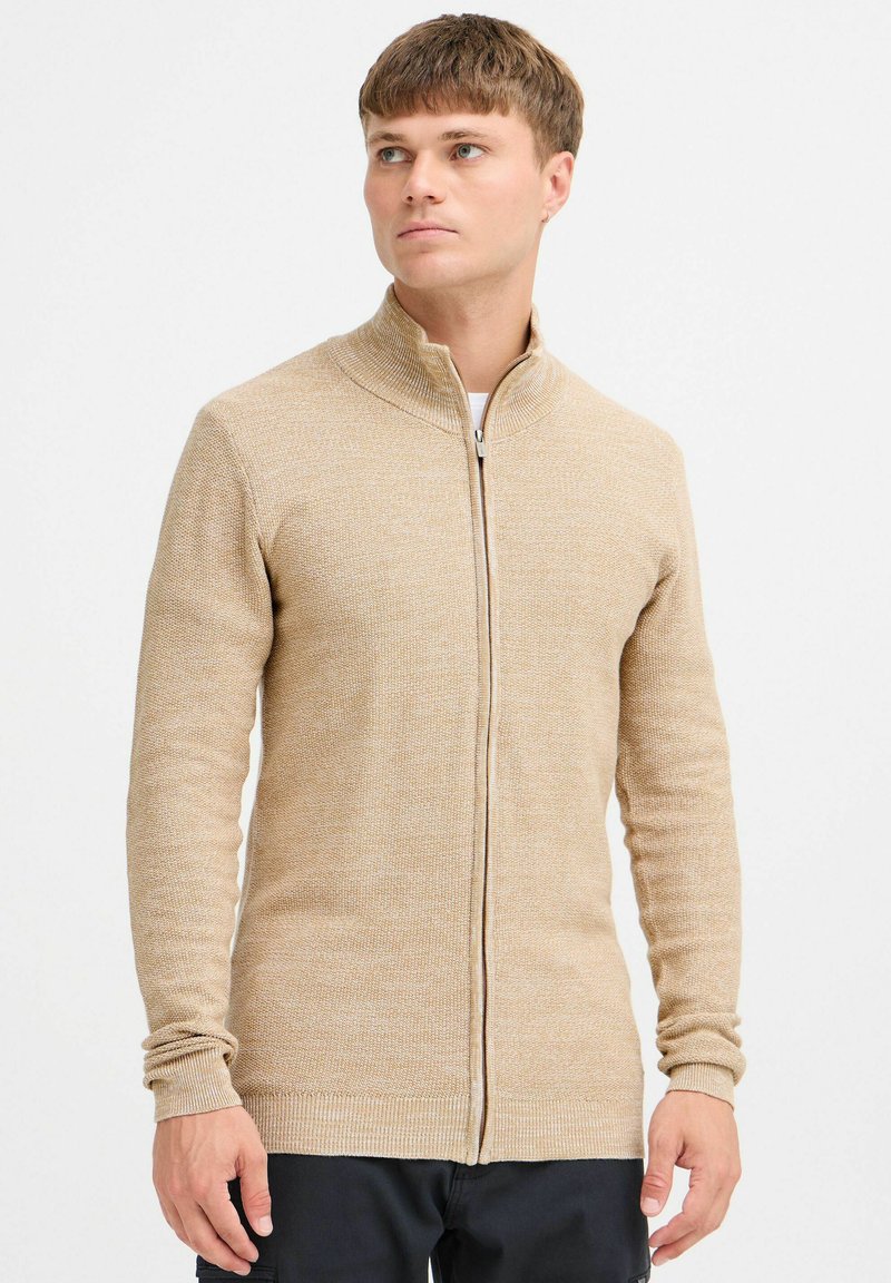 Sudadera cremosa con cremallera que presenta un cuello alto, mangas largas, un patrón de tejido texturizado y puños acanalados para un detalle adicional.