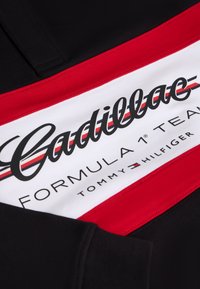 Vêtement noir avec un panneau rouge et blanc affichant la marque et le logo "Cadillac Formula 1 Team Tommy Hilfiger" en texte gras.