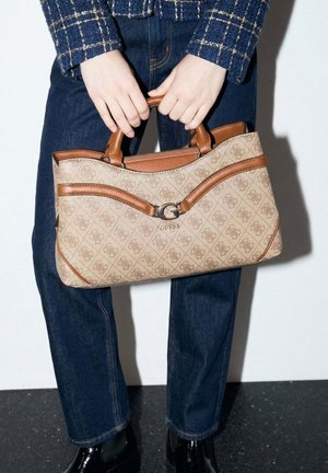 Mains tenant un sac à main beige à motifs Guess avec une garniture en cuir marron, porté avec un jean foncé, une veste à carreaux et des bottes en cuir verni noir.