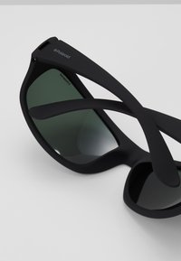 Ochelari de soare negri cu lentile verzi, având un design unic al ramei și brațe flexibile. Fabricati din material de origine biologică, cu o textură mată.