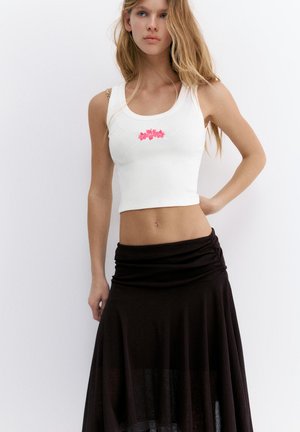 Junge Frau trägt ein weißes kurzes Tanktop mit pinkem Blumendesign und einem hoch taillierten schwarzen Rock, steht mit einer Hand in der Hüfte.