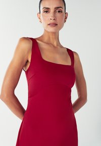 Červené šaty bez rukávov so štvorcovým výstrihom a priliehavým trupom, vyrobené z hladkej textílie. Dizajn obsahuje štruktúrované švy a elegantný siluetu.