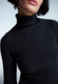 Črna rebrasta turtleneck jopica s tesnim krojem in teksturirano tkanino, ki prikazuje vertikalne vzorce in visok ovratnik.