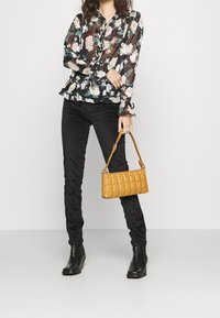 Blusa negra con estampado floral y dobladillo con volantes, botones en la parte delantera. Pantalones negros ajustados. Bolso acolchado amarillo con asa corta. Botines negros.