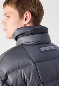 Jauns vīrietis profila skatā, ģērbts pelēkā, pildītā jaciņā ar augstu apkakli un redzamu Calvin Klein marķējumu uz muguras.