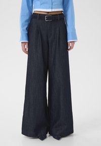 Pantalon en denim foncé à jambes larges avec une taille mi-haute, doté de passants de ceinture et d'une ceinture à boucle. Texture lisse et détails de couture minimaux.