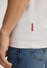 Gros plan sur une personne portant un t-shirt blanc avec une étiquette rouge "JOOP! JEANS" et un jean en denim bleu, montrant le bras et la main reposant sur le côté.
