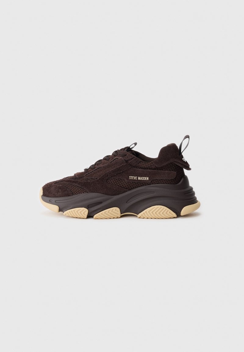 Zapatos deportivos marrones oscuros con una combinación de materiales de ante y malla. Suela de goma gruesa en color beige con un patrón texturizado y una lengüeta en el talón.