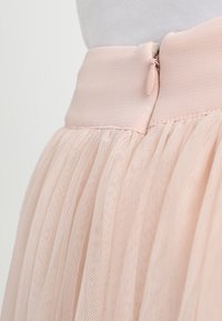 Jupe rose pâle en tulle texturé et plissé. La ceinture est lisse et comprend une fermeture éclair avec une petite languette.