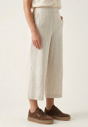 Vrouw draagt beige wijde cropped broek met zakken aan de zijkant en bruine suède sneakers met het label "Venezia" op een lichte achtergrond.