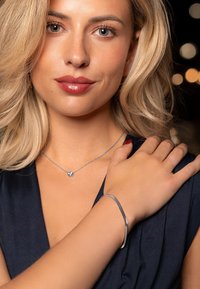 Bracelet argenté au poignet, collier délicat en chaîne argentée avec pendentif en forme de cœur, modèle aux cheveux blonds ondulés, portant un haut bleu foncé.