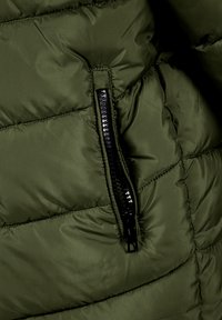 Section d'une doudoune vert olive mettant en avant une poche zippée noire, une texture matelassée et un tissu lisse avec des coutures accentuées visibles.