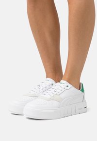 Hvide lædersneakers med en tekstureret overflade, grønne detaljer på hælen og tungen, rund tå og en tyk, rillede hvid sål.
