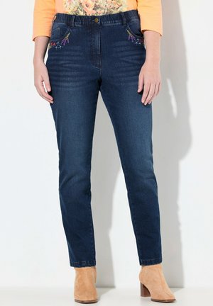 Femme portant un jean bleu foncé avec des broderies florales sur les poches, un haut à manches longues pêche et des bottines en daim beige à talons blocs.