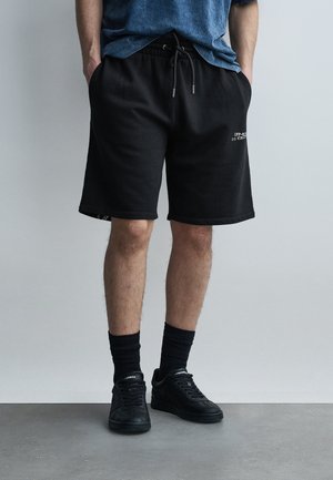 Άνδρας που φοράει μαύρα σόρτς με κορδόνι, μαύρες κάλτσες μέχρι τον αστράγαλο και μαύρα αθλητικά παπούτσια, στέκεται μπροστά σε απλό φωτεινό γκρι φόντο.